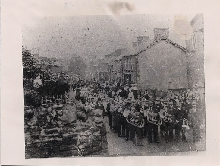 Members of Ystalyfera Public Band marching playing instruments with people watching